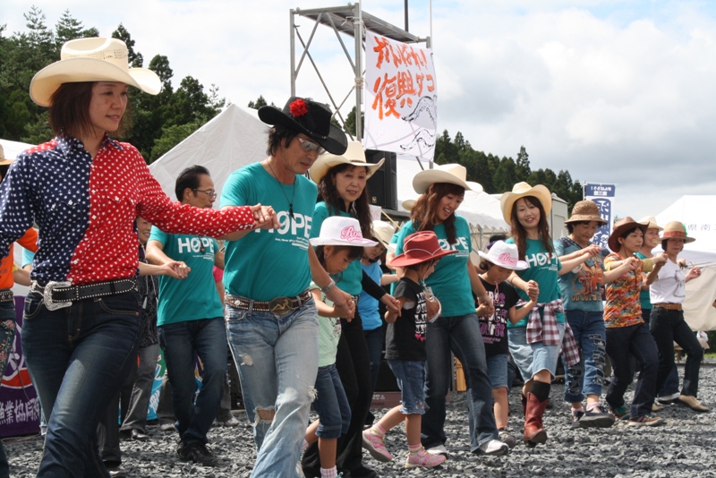 カウボーイハットをかぶった大人や子どもたちが手をつなぎ、笑顔でダンスを楽しんでいる様子の写真