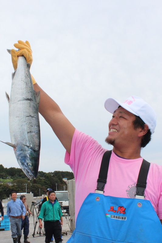 ピンク色のTシャツと青い胸当て付きズボンを着た男性が、大きな秋サケを片手で高く掲げ、笑顔で見せている様子の写真