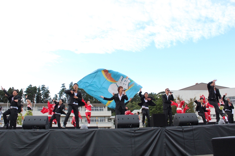 ステージ上でEXILE（エグザイル）のメンバーがマイクを持って歌いながらパフォーマンスをし、後ろでは赤や黄緑の衣装を着た人たちが踊り、大きな青い旗が振られステージが盛り上がっている様子の写真