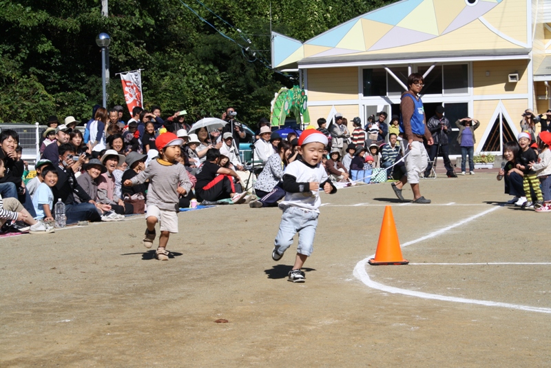 園庭で赤い帽子をかぶった2人の園児がオレンジ色のコーンを目印に走り、その様子を大人たちが日傘やカメラを手に笑顔で応援している競走の写真