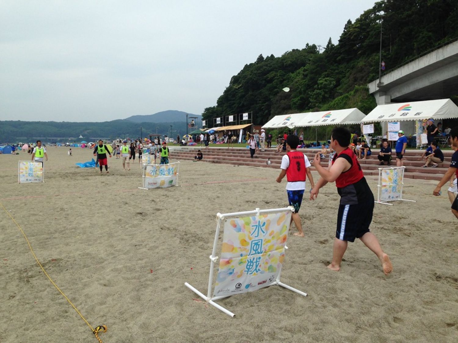 砂浜に設けられたコートで赤と緑のビブスを着た参加者たちが水風船を投げ合いながら対戦し、楽しんでいる様子の写真