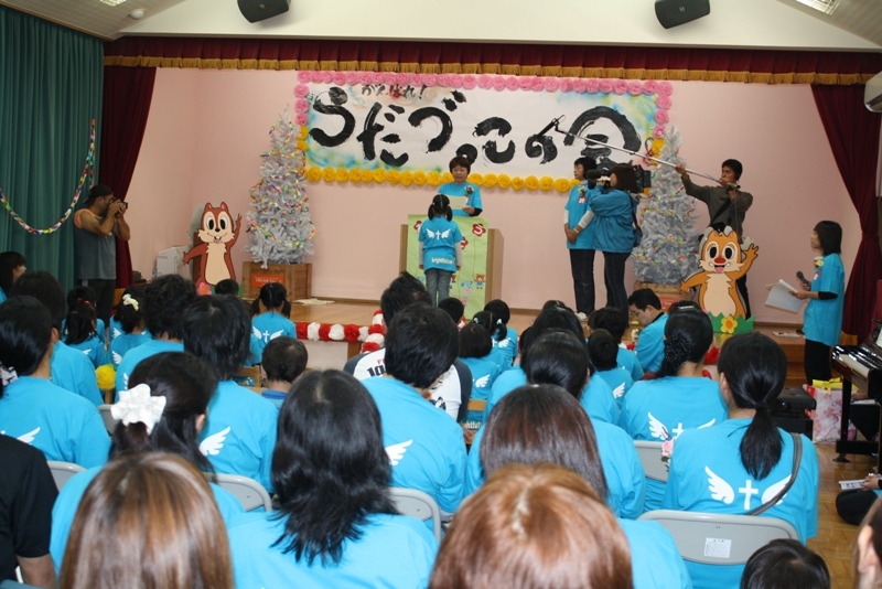 青い揃いのシャツを着た参加者たちが見守る中、正面の演台では同じ青いTシャツを着た子どもが『がんばったで証』を受け取り、周囲のスタッフやカメラマンが見守る中で授与式が行われている様子の写真