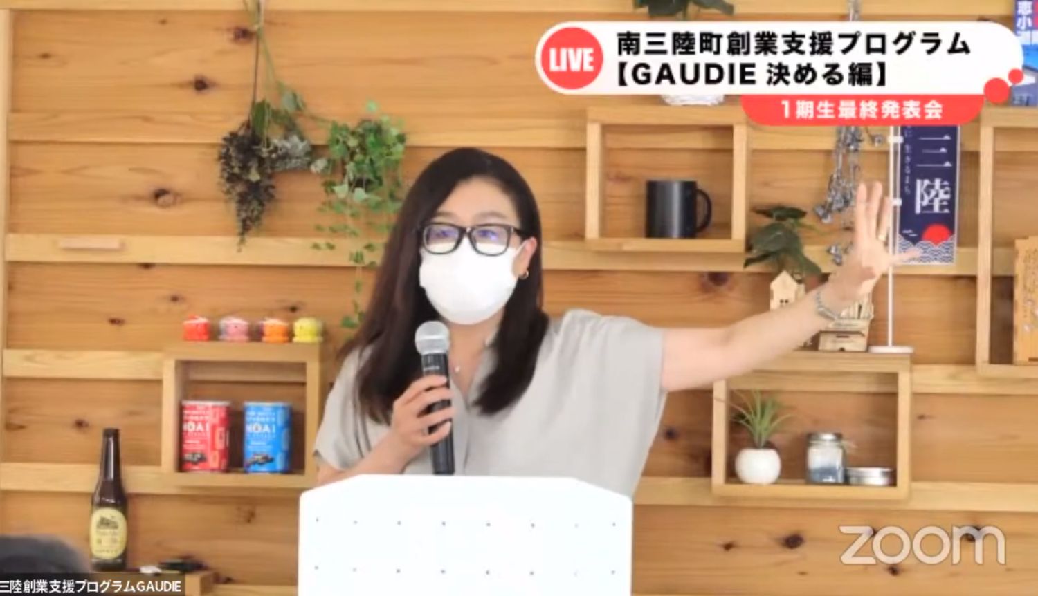 眼鏡をかけた女性がマスク姿で右手を大きく上げながらマイクを持ち、南三陸町創業支援プログラムGAUDIEの発表会でスピーチをしている様子の写真