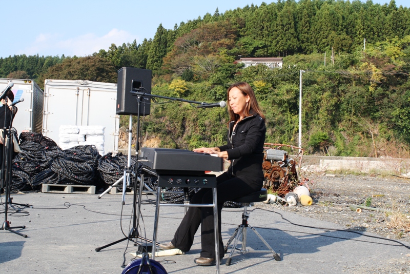 晴天の中、屋外の会場で八神純子さんがキーボードを弾きながら歌を披露しているチャリティーライブの様子の写真