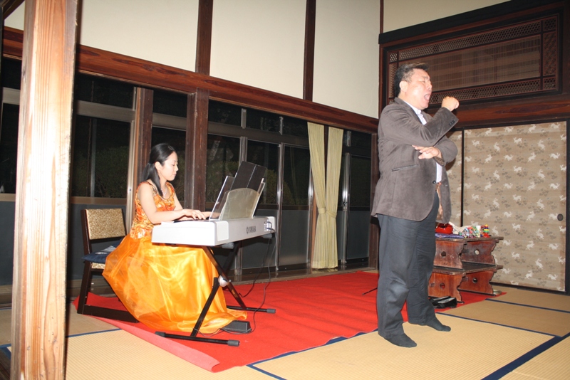 興福寺の会場で、オレンジ色のドレスを着た田中奈織美さんが電子ピアノを弾き、その横で池田弦さんがマイクを手に歌っているお月見コンサートの様子の写真