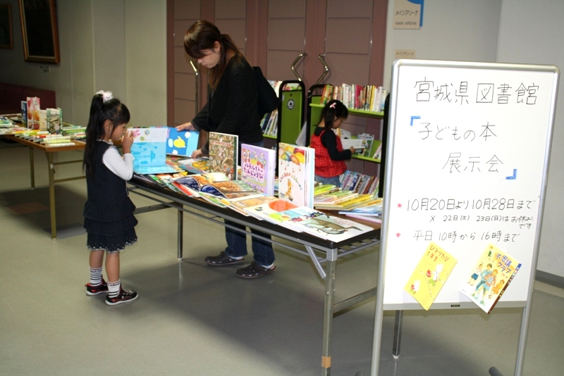 展示会の案内が書かれたホワイトボードが立てかけられた会場で、長机に絵本や児童書が見やすく並べられ、女性が本を手に取り、女の子がページをめくって見ており、奥の棚でも別の子どもが本を手にしている子どもの本展示会の写真