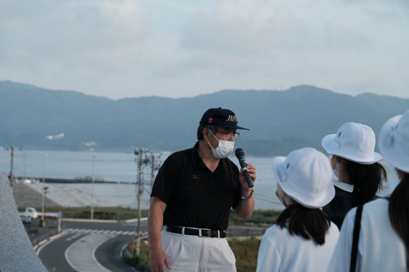 帽子をかぶった佐藤誠悦さんが、マイクを使って白い帽子をかぶった女子学生たちに向けて説明をしている様子の写真