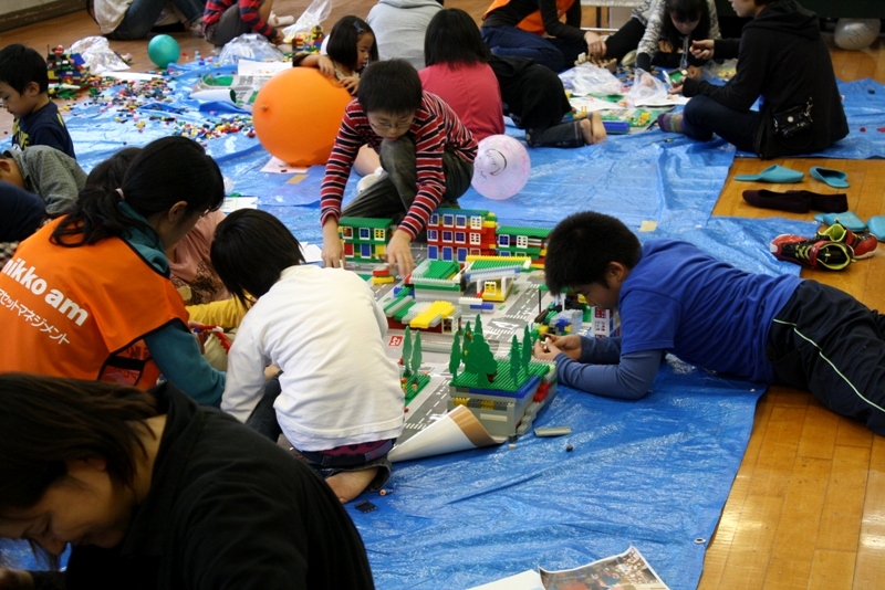 室内の床にブルーシートが広げられ、その上で子どもたちがカラフルなブロックを使って建物や道路などを組み立てながら街づくりを楽しんでいる様子の写真