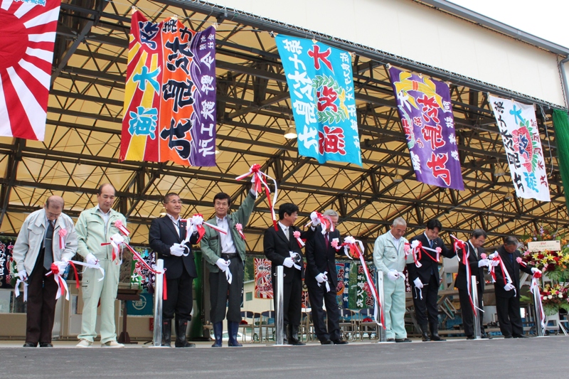 仮設魚市場のオープンセレモニーで、色鮮やな大漁旗が掲げられる中、関係者が並んでテープカットを行っている様子の写真