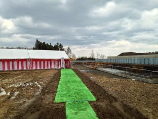 土の地面に安全祈願祭会場の紅白幕のテントへと促す、緑色の人工芝で作られた大きな矢印が伸びている、広々とした建設予定地の空き地の写真