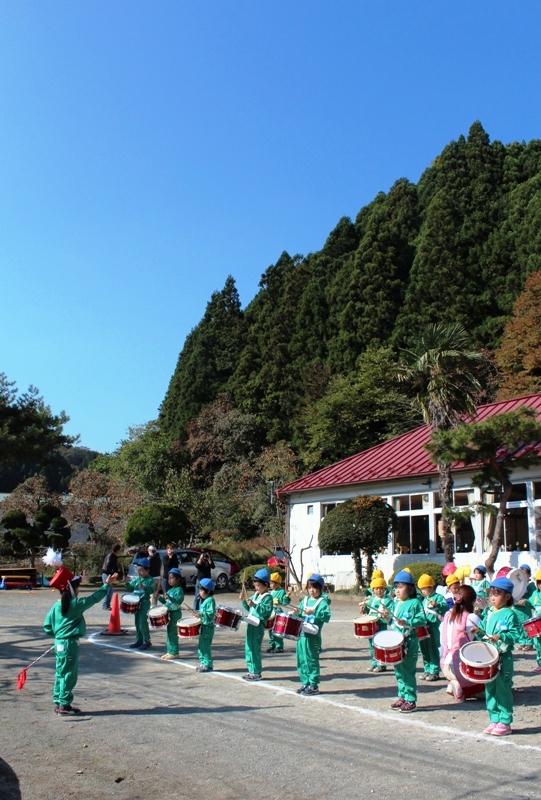 園庭で、あさひ幼稍園の園児たちが緑の体操服を着て鼓笛隊として整列し、太鼓を叩いて演奏している様子の写真