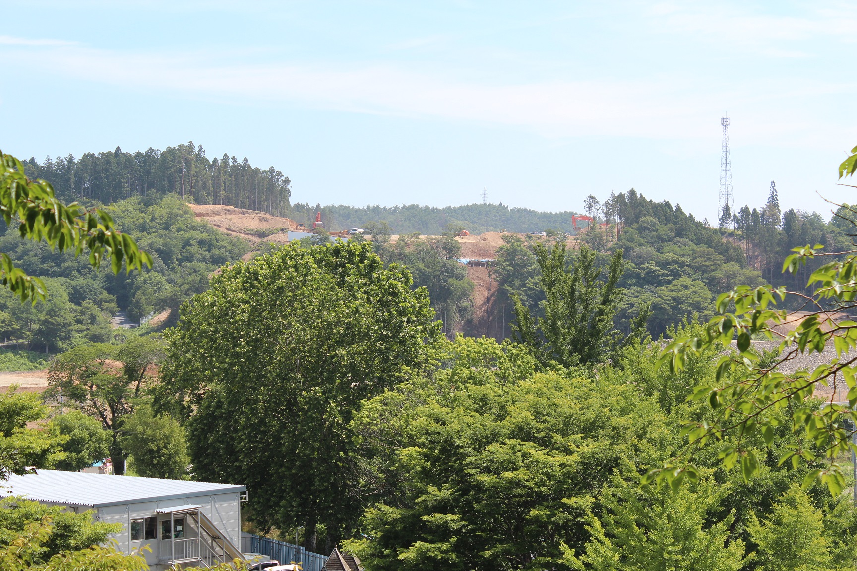 手前左側に外付け階段のあるプレハブ小屋があり、周りに青々とした木が生え、奥の切り開かれた造成地に複数の重機が見える写真
