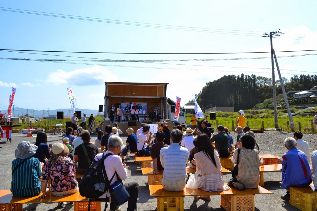 青空の下の即席ステージで、虹色イラストコンクールの授賞式が行われている様子を、たくさんの人がベンチに座って見ている写真