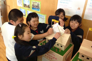 小学生の子どもたち5人が、屋根付きの小さな家の形をした「書き損じはがき回収ポスト」に楽しそうにハガキを投函している様子の写真