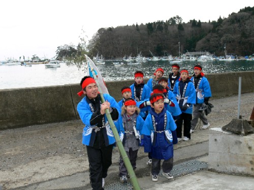 防波堤のような場所で、青い法被を着て赤い鉢巻をした数人の子供たちが、旗をつけた大きな青竹を担ぎながら、歩いている様子の写真