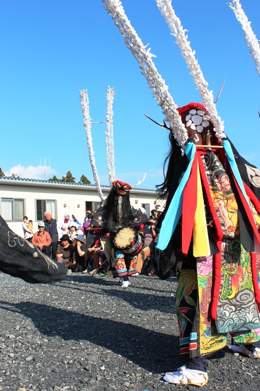 色鮮やかな装束を身に着けた踊り手たちが太鼓を打ちながら鹿子躍を披露し、観客が見守っている様子の写真