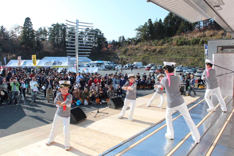 ステージ上で水兵の衣装を着た子どもと大人の出演者たちが踊りを披露し、会場前には多くの観客が集まって見守っている福興市の様子の写真