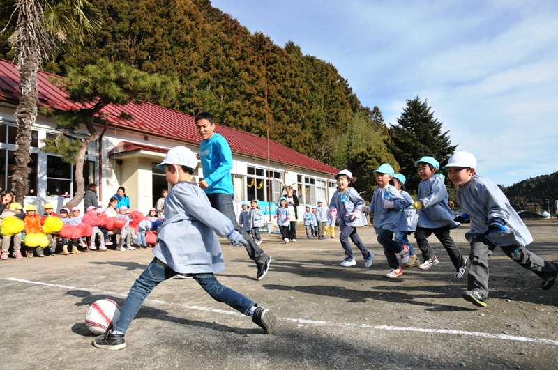 長谷部選手が園児たちとグラウンドでサッカーを楽しみ、ポンポンを持って応援する子どもたちの姿も見えるサッカー交流の様子の写真