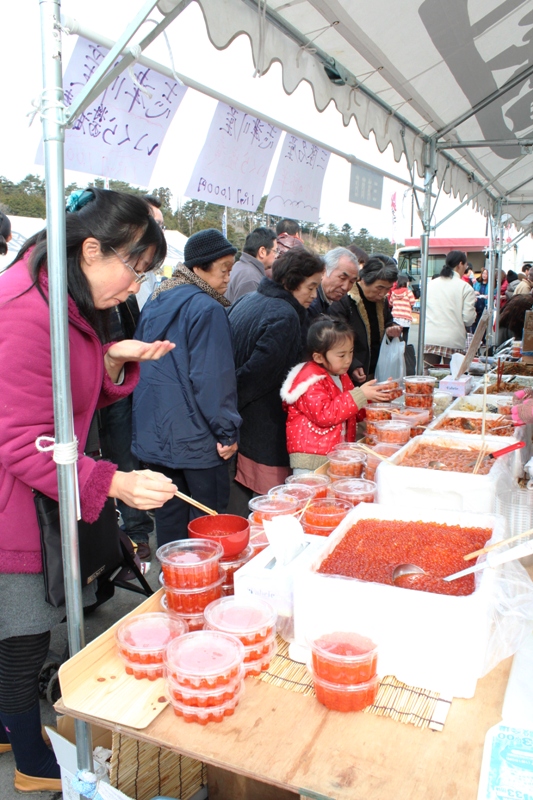 いくらや魚介類などが並ぶ屋台の前で、多くの来場者が試食したり品定めをしているおすばで福興市の様子の写真