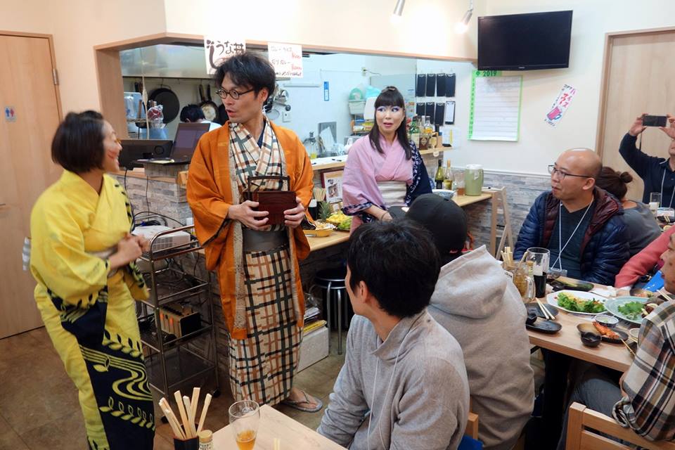 テーブルの上に食事や飲み物が置かれた飲食店の店内で、浴衣姿の3人が立って演技をし、座っている観客が見守っている写真