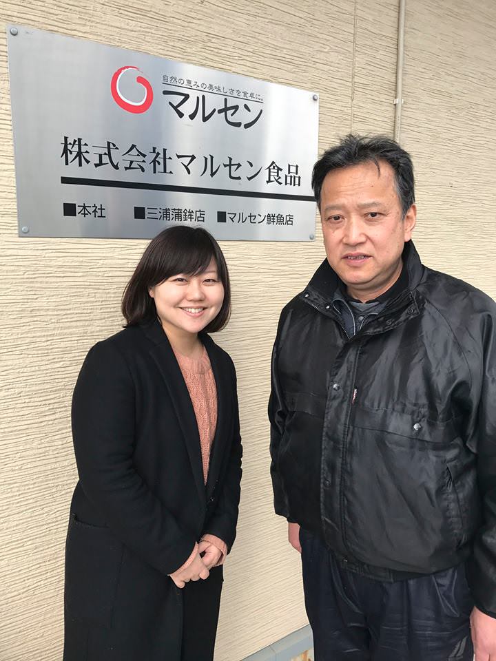 「株式会社マルセン食品」と書かれたシルバーの看板が設置された壁の前に並んで立っている女性リポーターと黒いジャンバーを着た男性の写真