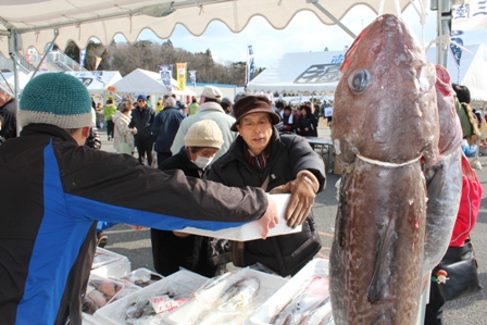 屋外のテントの下で開催された魚まつりの出店で、来場者が大きな魚を前に販売員から商品を受け取っている様子の写真