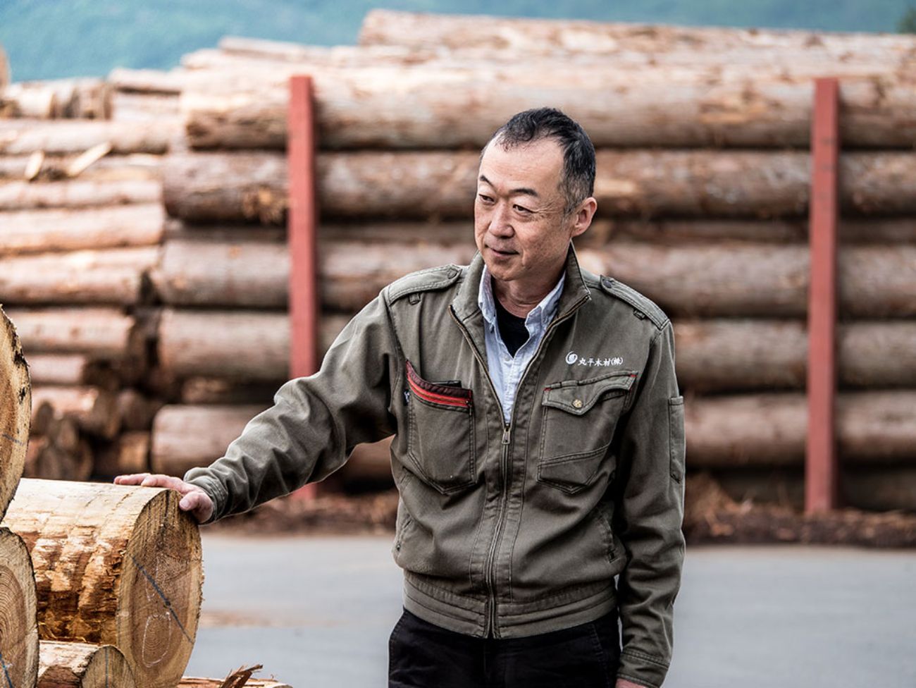 積み上げられた丸太の前で作業服姿の小野寺邦夫さんが木材に手を置き立っている写真