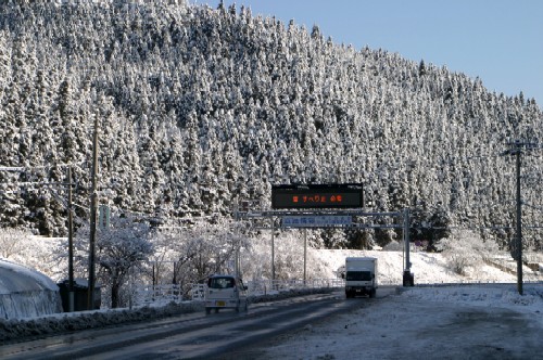 針葉樹の森が一面に雪を被り白く覆われた山を背景に、雪の残る道路の上を車が行き交い、道路上には「雪すべり注意」と表示された電光掲示板が設置されている冬の風景写真