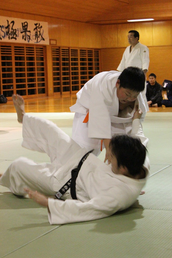 道場の畳の上で少年が黒帯の先生に向かって投げ技をかけ、先生が受け身を取っている練習の様子を写した写真