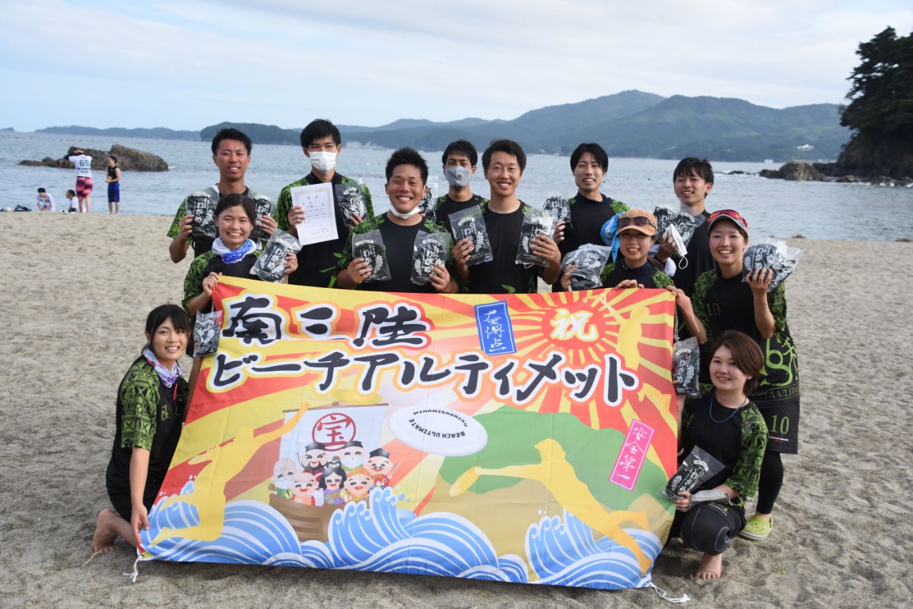 砂浜で黒と緑のユニフォームを着たビーチアルティメットのチームメンバーが、「南三陸ビーチアルティメット」と書かれたカラフルな横断幕を前に笑顔で並び、手には賞品と思われる黒い袋や賞状を持ちながら、後ろには穏やかな海と山々が広がっているビーチアルティメット大会の表彰風景の写真