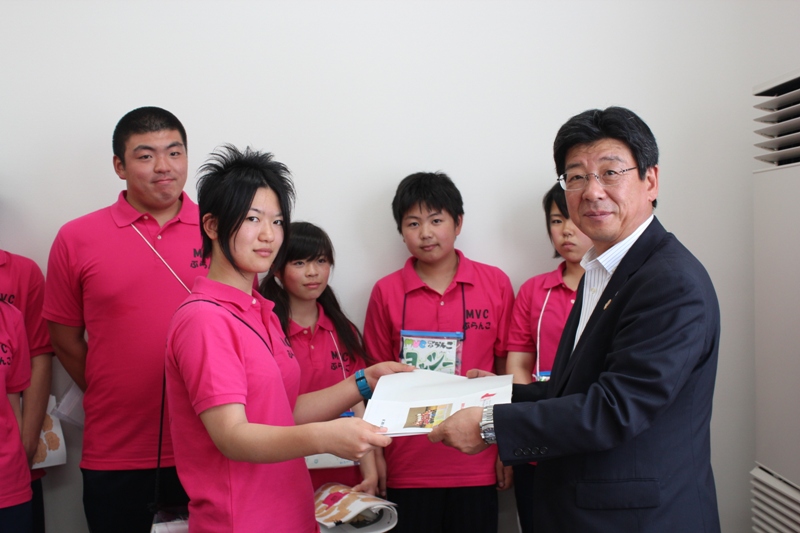 ピンクのポロシャシャインを着たジュニア・リーダーの皆さんが集まり、代表の一人が、町長に提案書を手渡ししている様子の写真