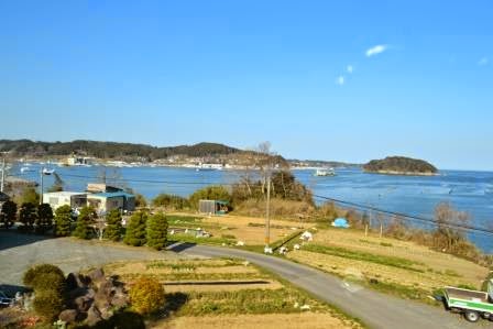 青空の下、海沿いに緑に囲まれた家々が並び、遠くには船が航行する水面が見え、手前には道路と畑のような耕地が広がる、穏やかな海辺の風景を写した写真