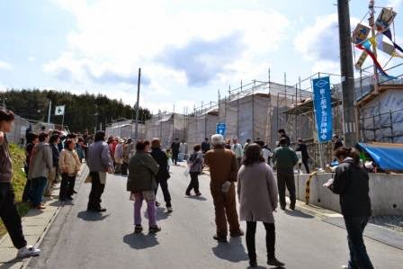 道路に集まった多くの地域住民が、傾斜のある道路沿いに立つ建設中の家を見上げながら上棟式の開始を待っている写真