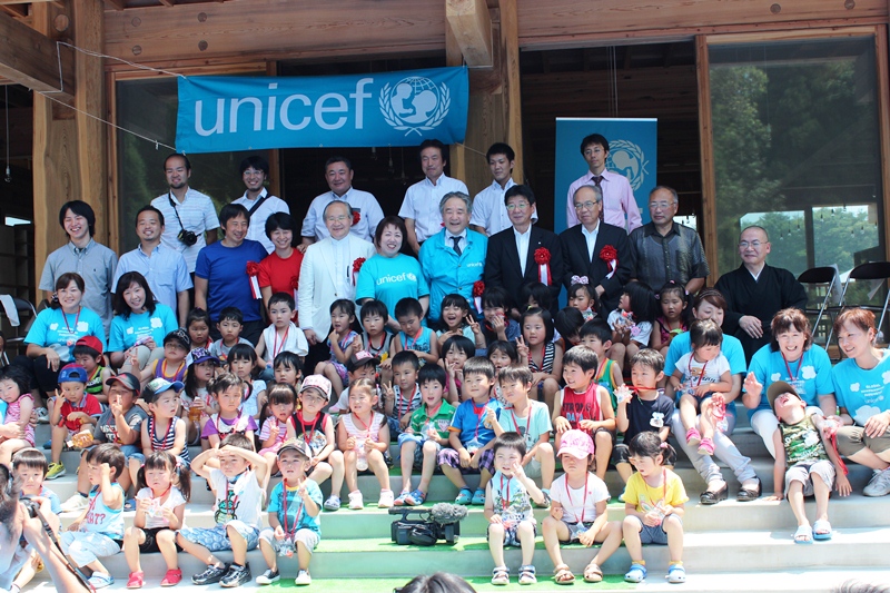 木造の園舎に水色のユニセフの旗が掲げられ、たくさんの子どもたちと関係者が笑顔で記念撮影に収まっている様子の写真