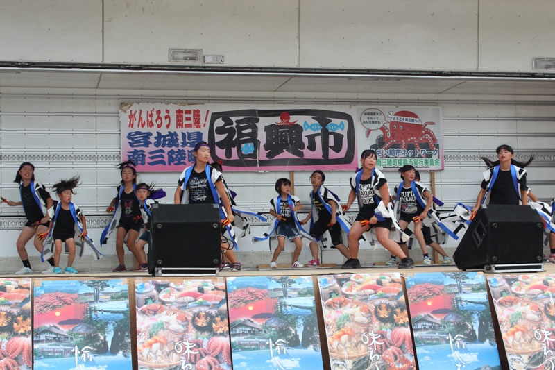 「がんばろう 南三陸！復興市」と書かれたステージで、、法被を着た子どもたちが元気いっぱいに踊りを披題している復興市のイベントの様子の写真