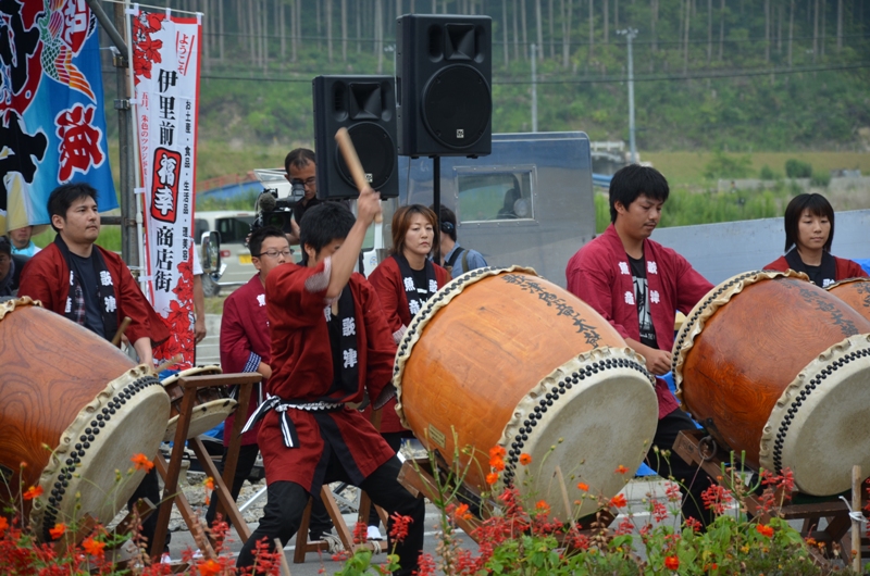 赤い法被を魚竜太鼓の方々が野外のステージで力強く太鼓を打ち鳴らし、観客に向けて迫力ある演奏を披題している、地域の祭りの太鼓演奏の様子の写真