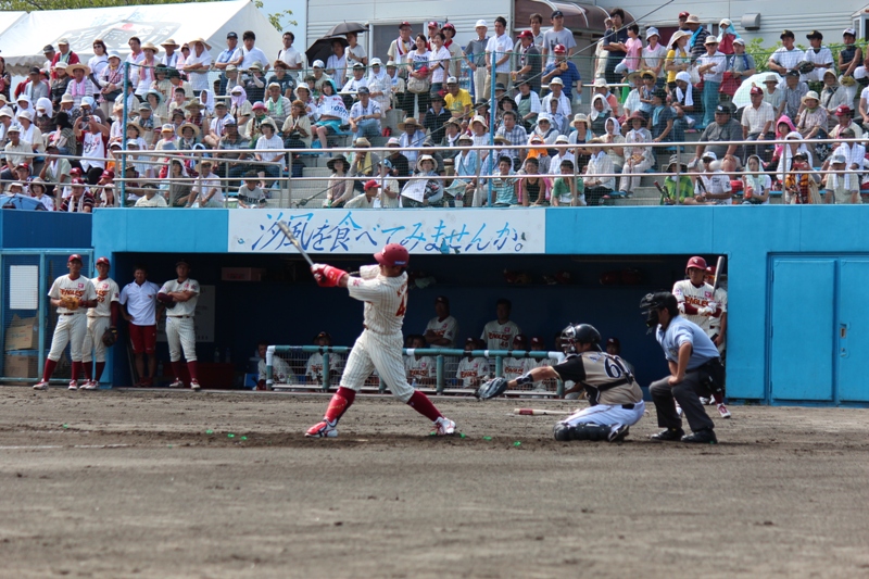 満員の観客が見守る球場で南三陸町復興支援試合が行われ、打者が豪快にスイングし、捕手と審判が構えるプレー中の様子の写真