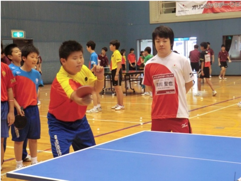 体育館の中で卓球台を囲み、赤と黄色のユニフォームを着た少年が真剣な表情でラケットを振り、そのそばで岸川選手がフォームを見守りながら指導している卓球教室の様子の写真