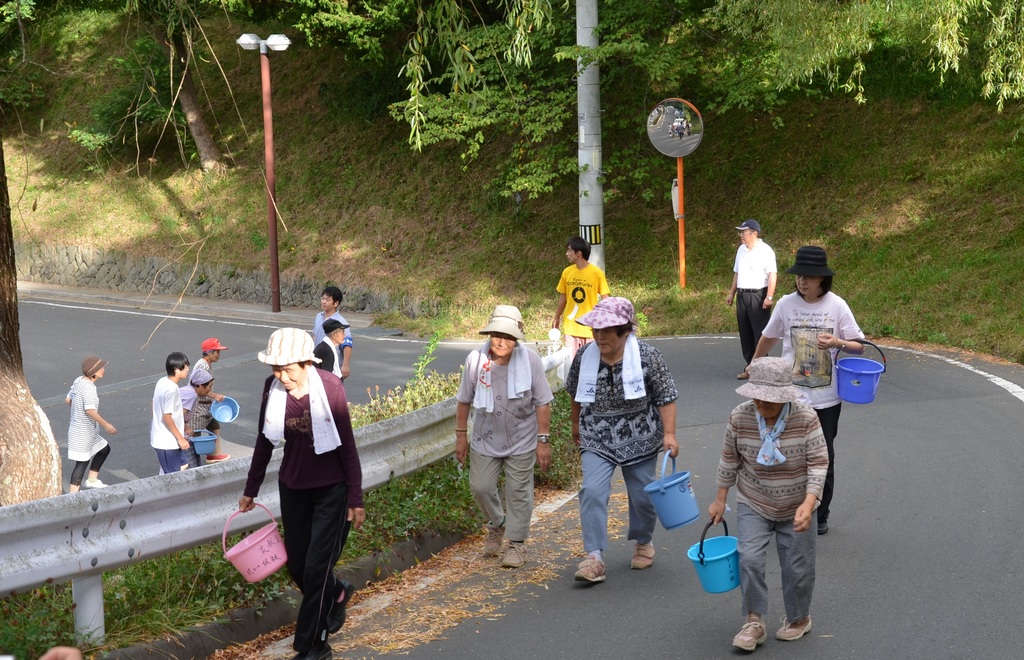 坂道を上る避難路で、帽子をかぶった高齢者や地域の人たちがバケツを手に持ち、タオルを首にかけながら真剣な表情で歩いており、後方には若い人たちの姿も見える地震・津波防災訓練の様子の写真