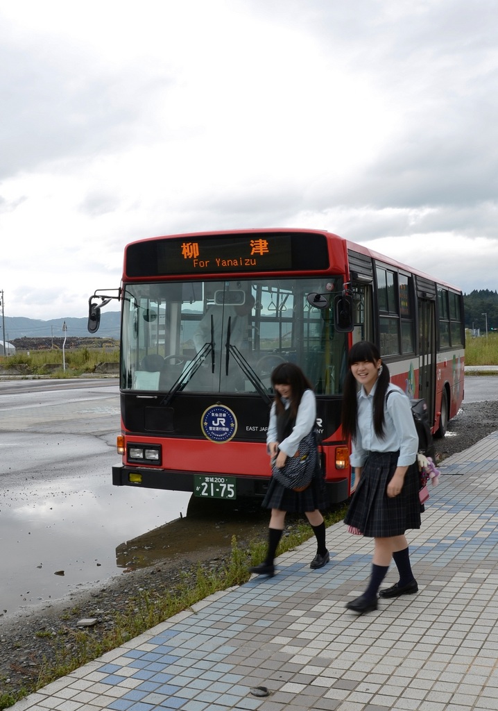 赤色の柳津行きのBRT専用バスが停車しており、バスの前で制服姿の女子高校生たちが笑顔で歩いている様子の写真