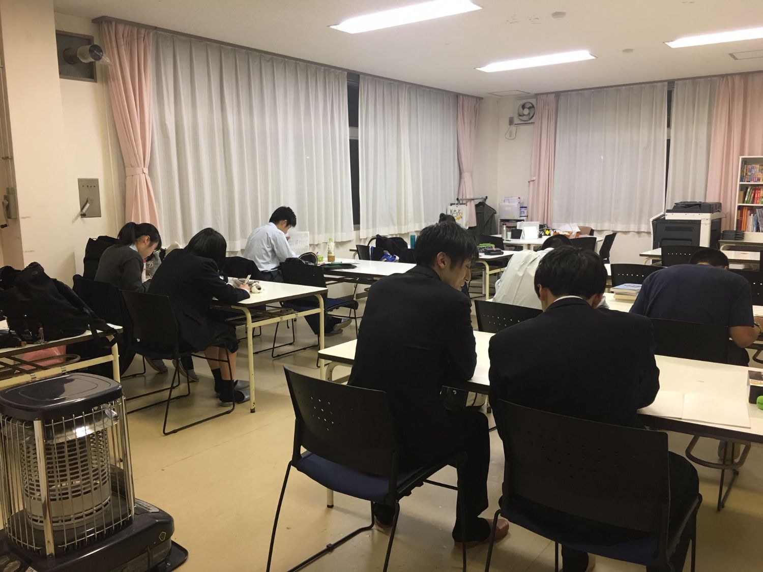 壁際にはカーテンのかかった窓と本棚が設置された教室内で、数人の生徒と男性が机の席に座り、それぞれ勉強している様子の写真