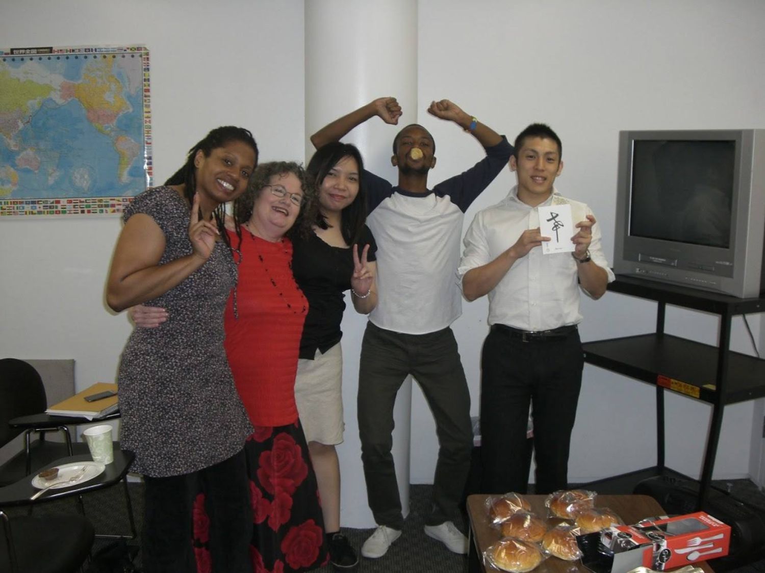 地図やテレビが置かれている室内で、1人の日本人と4人の男女が楽しそうに並び、中央の男性は口にパンをくわえて両手を上げてポーズを取り、他の人たちは笑顔でピースをしている写真