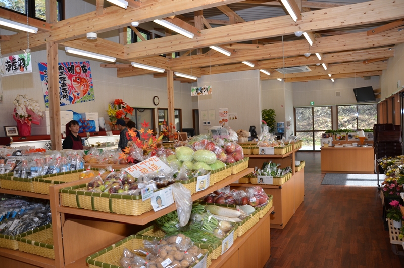キャベツ、りんご、じゃがいも、にんじん、葉物野菜などの生鮮食品が棚や平台に並び、店員がカウンターで接客している直売所「みなさん館」の店内の様子の写真