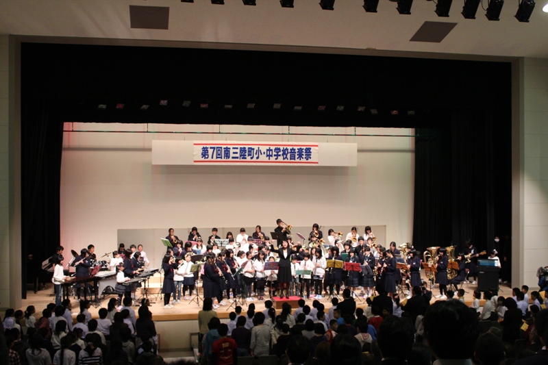 第7回南三陸町小・中学校音楽祭の横断幕が掛かるホールのステージ上で、数十人の小学生と中学生が並び、指揮者が中央で腕を振って合奏を指揮し、トランペットやトロンボーンなどの金管、フルートやクラリネットなどの木管、打楽器やなど多様な楽器が演奏され、客席には保護者や観客が大勢詰めかけている写真