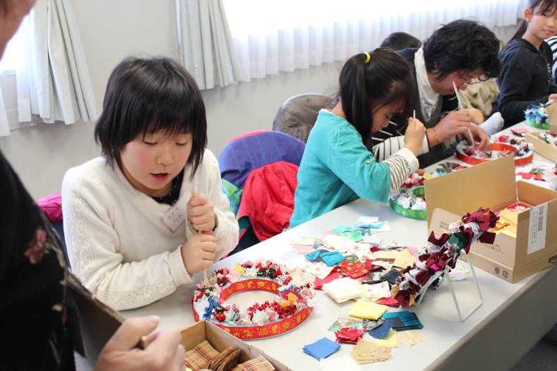 子どもたちがテーブルに並んで座り、赤や緑など色とりどりの布や紙片、リボンを使って円形の台に飾りを差し込みながら真剣にクリスマスリースを作っている様子を写した写真
