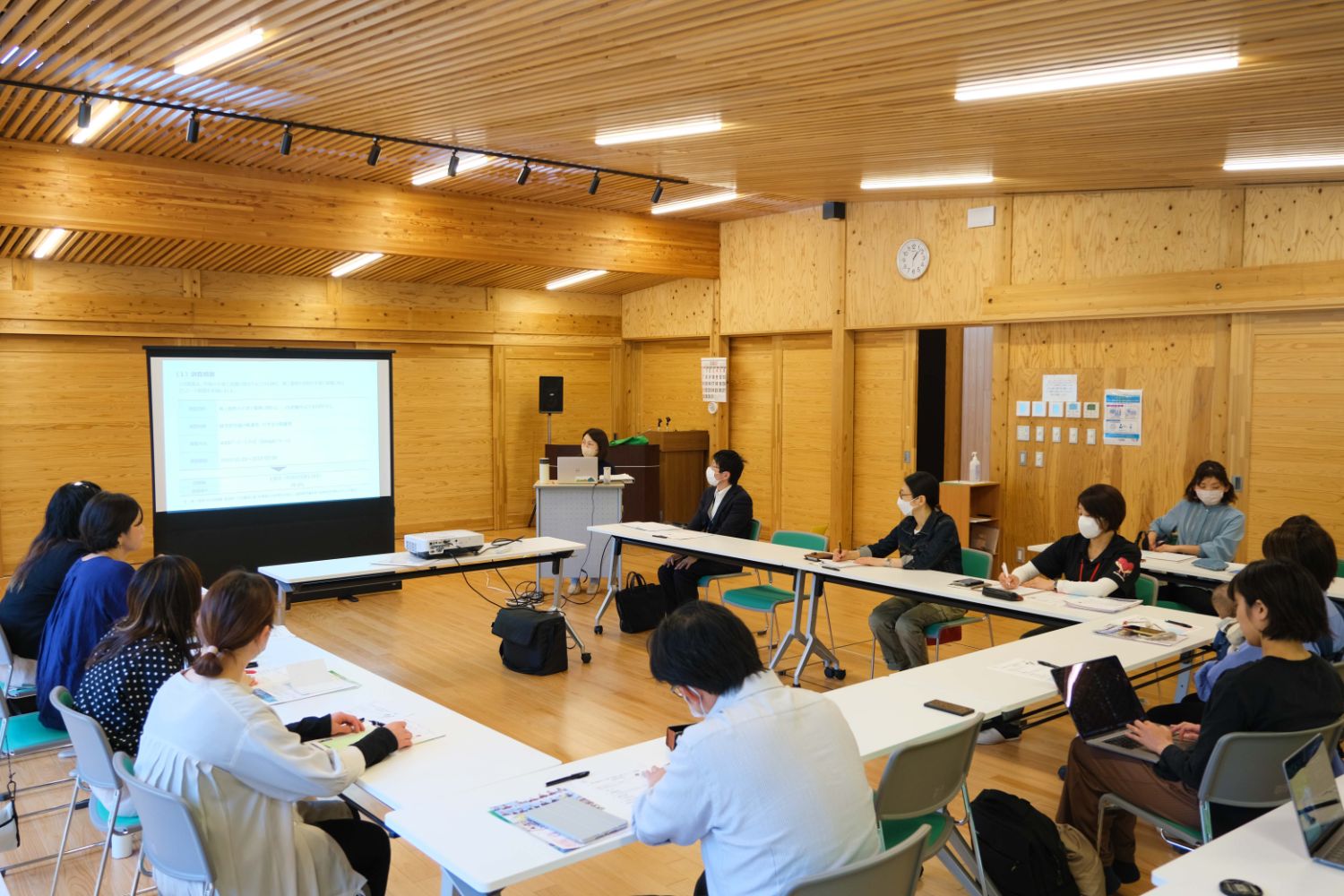 席に座った参加者たちが前方のスクリーンに映った資料に注目している様子の写真