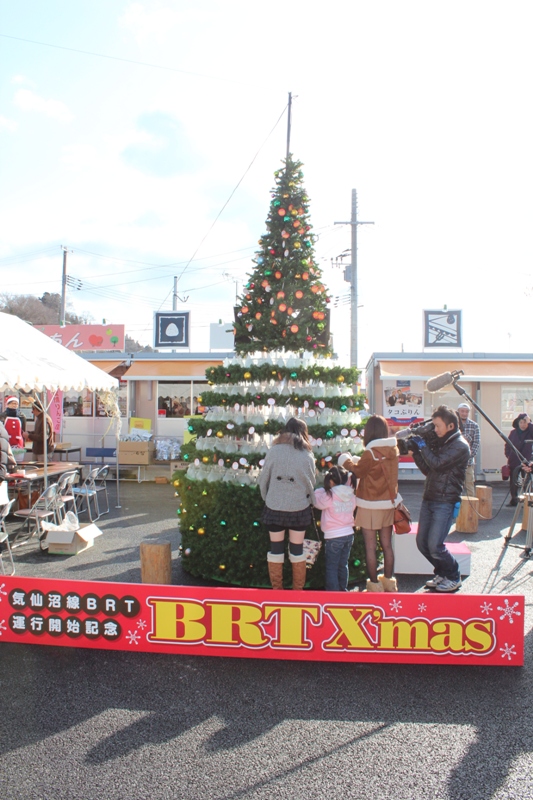 屋外に設置されたクリスマスツリーの前で、訪れた女性と女の子が飾りつけを行い、テレビカメラがその様子を撮影しているJR気仙沼線BRT運行開始記念イベントの様子を写した写真