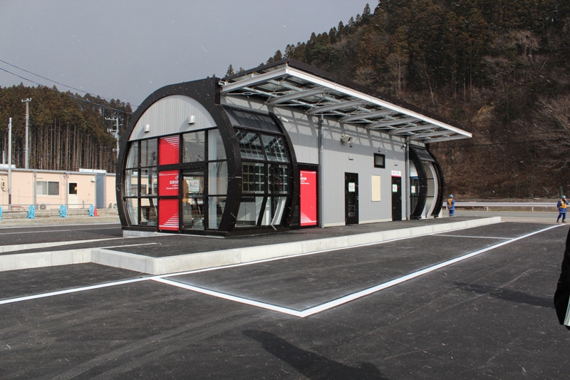 黒いアーチ状の骨組みと大きなガラス窓を組み合わせた近代的な小型の駅舎が立ち、入口に赤い扉と庇が設けられ、背後に緑の木々と山が見える場所に立つJR志津川駅の外観写真