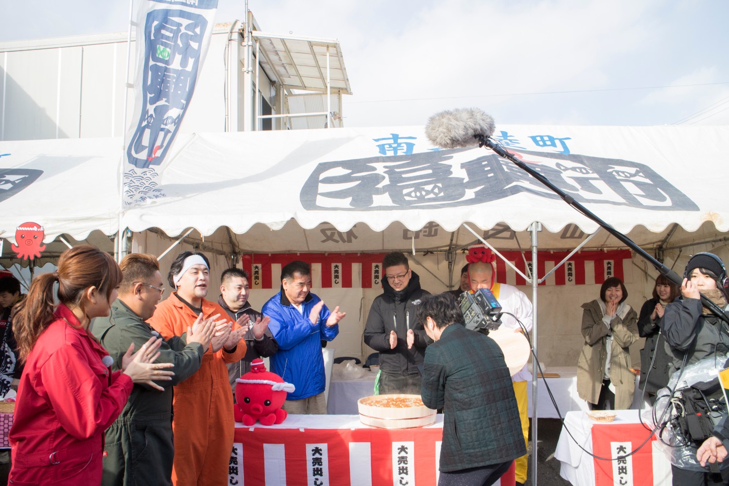 「南三陸町福興市」と書かれた白いテントの前で、サンドウィッチマンさんと大勢のスタッフや参加者が拍手をして囲み、中央ではテレビクルーが巨大な海鮮ちらし寿司を撮影している様子を捉えた福興市のイベントの様子を写した写真