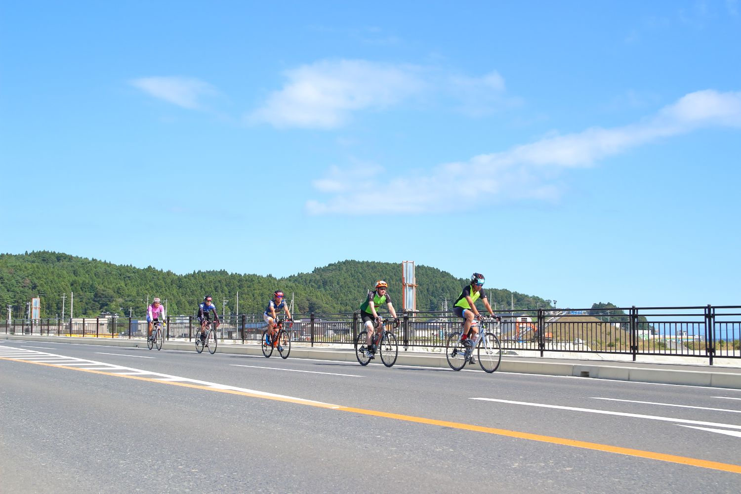 海が見渡せる景色の良い道路上を、縦一列に並んで自転車を漕いでいる5人のライダーの方々の写真